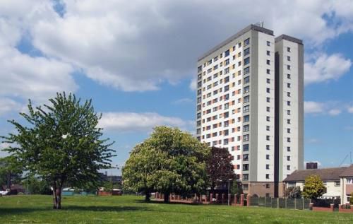 A view of an 18 storey tower block next to a house with a park of grass and trees in the foreground.